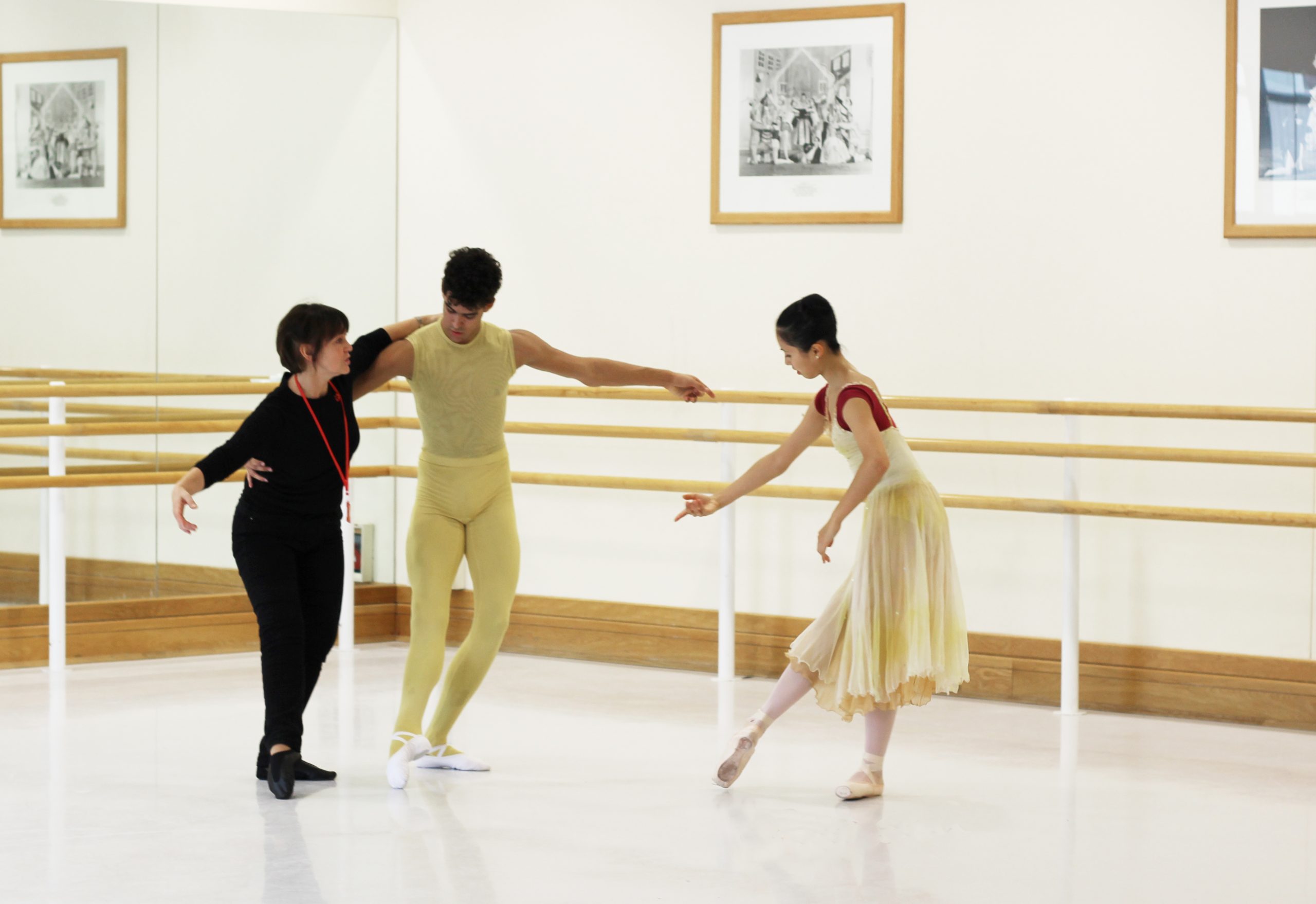 Leslie collier wears a long-sleeve black shirt, black trousers and black shoes in the studio. She holds the back of a male student with her left arm. The student wears a yellow short sleeve top and yellow tights with white shoes. Both tendu their right legs forward in plie on the left leg. A female dancer stands to their left, tenduing in the same pose towards them. She wears a red cap sleeve leotard and a yellow sleeveless dress on top. She has pink tights and pointe shoes.