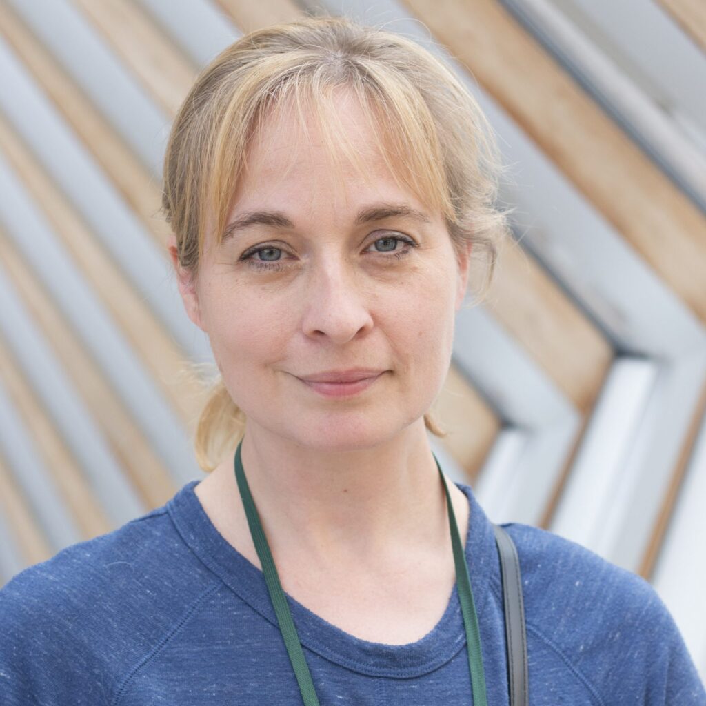 Sarah smiles at the camera on the bridge of aspiration with her blonde hair tied back in a ponytail. She wears a blue crew neck shirt with a green lanyard around her neck.