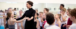 The royal ballet school launches enlighten teacher training webinars A teacher demonstrating with a student surrounded by a group of young dancers.