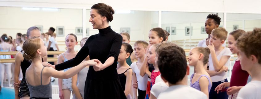 Enlighten A teacher demonstrating with a student surrounded by a group of young dancers.