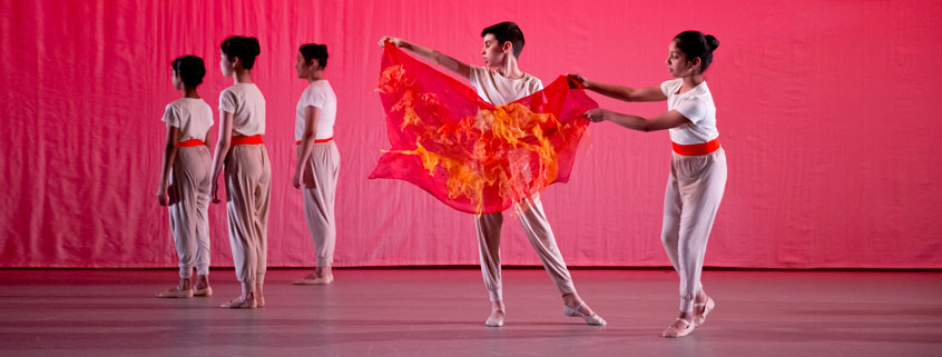 Five primary steps students performing on a stage, all wearing white, dancing in front of a pink background holding a orange piece of fabric.