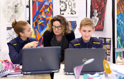 Two students at their laptops in an art class, with their teacher reading their screens over their shoulders.