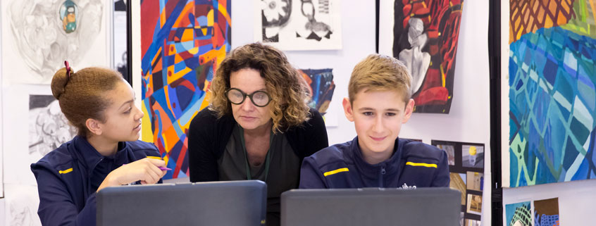 Two students at their laptops in an art class, with their teacher reading their screens over their shoulders.