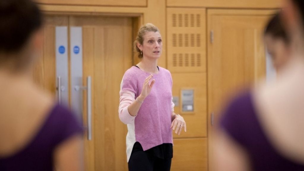 Ballet coach zenaida janowsky stands in front of a group of students speaking to them.