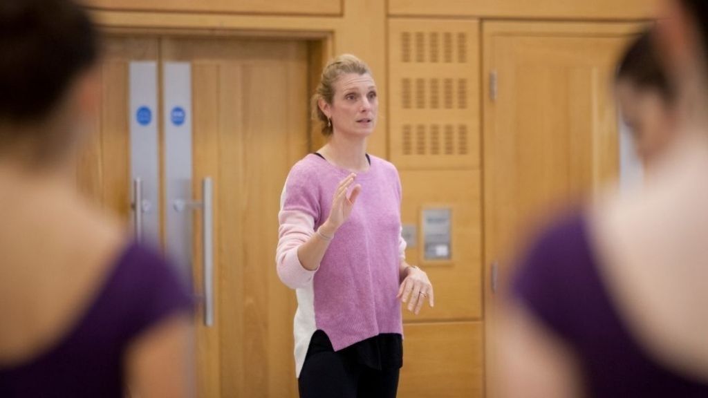 Ballet coach zenaida janowsky stands in front of a group of students speaking to them.