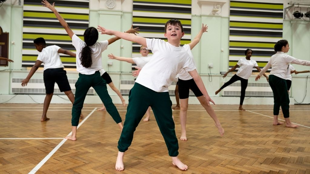 A group of children in white t-shirts and green trousers dance with their arms stretched out to the side or in the air