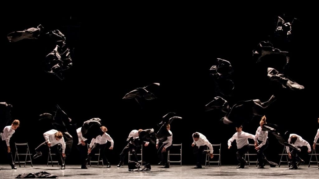 A group of dancers on stage in a semi circle take off their black blazers and throw them in the air