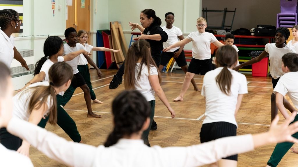 A group of primary steps students dance around a sports hall with their teacher in the middle