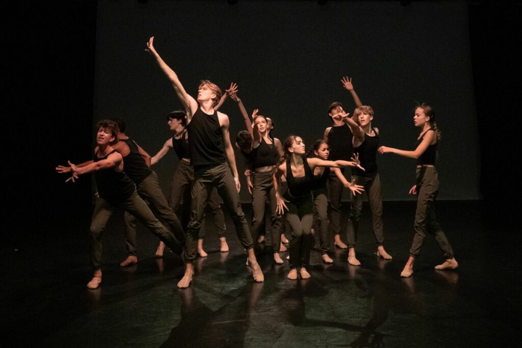 A collective of dancers weaving in and out of each other reaching up and down, dressed in green trousers and black tops.