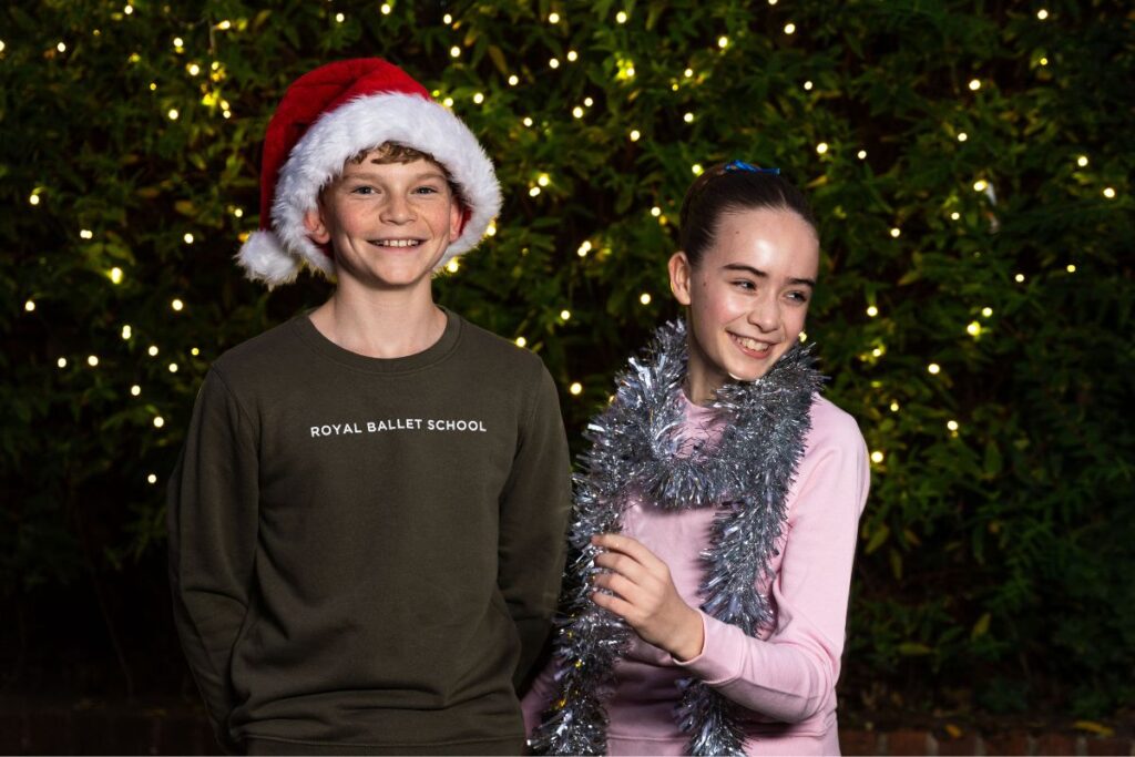 Two students in front of a tree with lights, wearing royal ballet school jumpers, tinsel and santa hats.