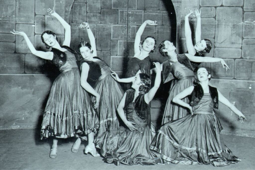 Ninette de valois as a young dancer surrounded by other dancers in long skirts and posing in different dance positions.