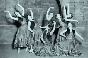 Ninette de valois as a young dancer surrounded by other dancers in long skirts and posing in different dance positions.