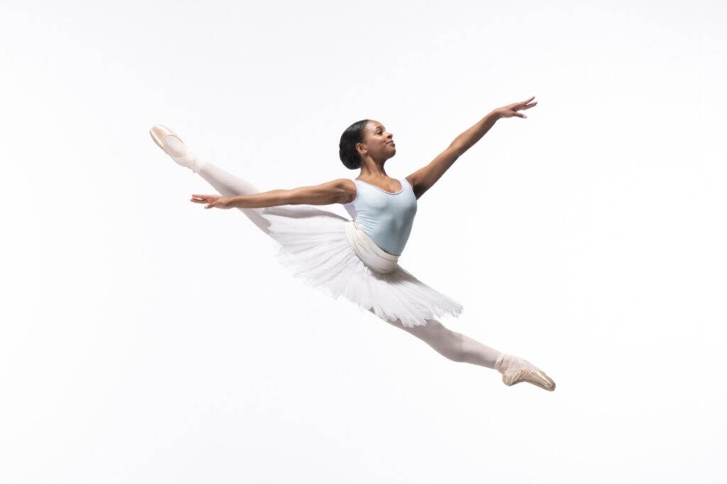 Rebecca myles stewart in a light blue leotard and white tutu in a sissone jump.