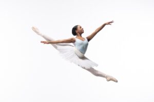 Rebecca myles stewart in a light blue leotard and white tutu in a sissone jump.