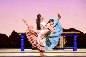 Alum and principal of the royal ballet francesca hayward in a long blue dress kicking her leg forward pictured with year 8 student ariella wearing a pink dress kicking her leg to the back.