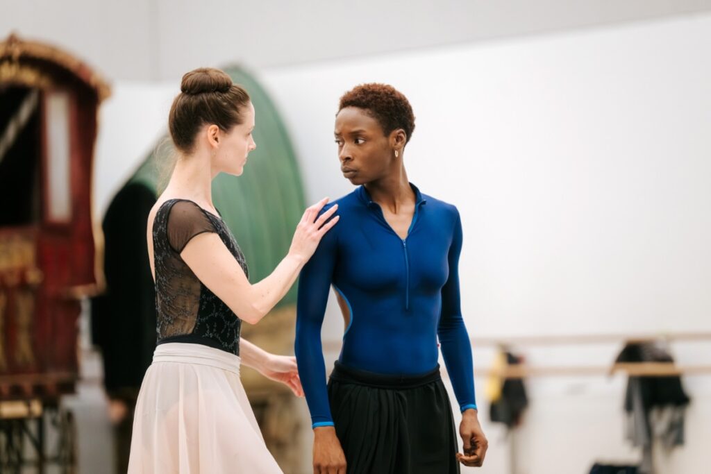 Rishan in a blue long-sleeve leotard and short hair looking intently at a fellow dancer in a black leotard, light pink skirt and brown hair in a bun