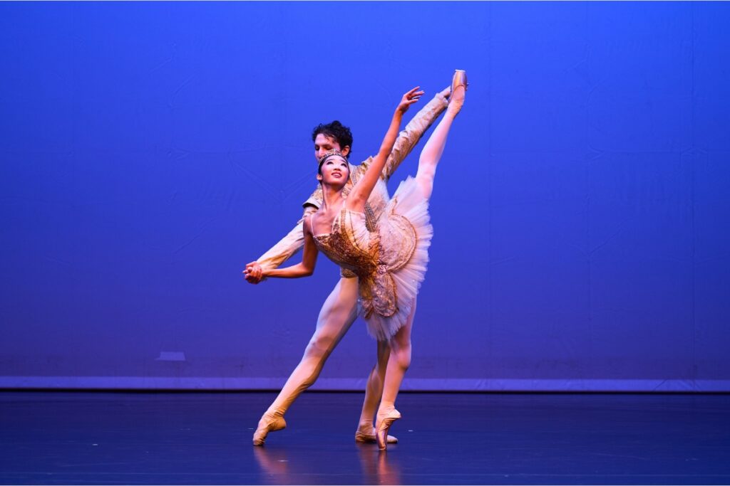 Julian and sophia pose in grand pas classique. Julian tendus to the side with his right leg while holding sophia's hand. Sophia is in an arabesque penche holding julian's right hand with her right. She looks up at her left hand.