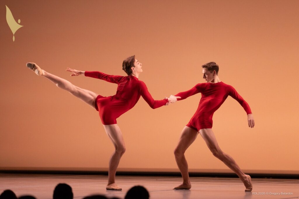 Henry dressed in a red long sleeve unitard with shorts partners another male dancer. Henry, right, leans back with his legs bent while holding his partner's hand. His partner is in an arabesque facing henry, left leg extended and right leg bent. His left arm reaches back in line with his leg.