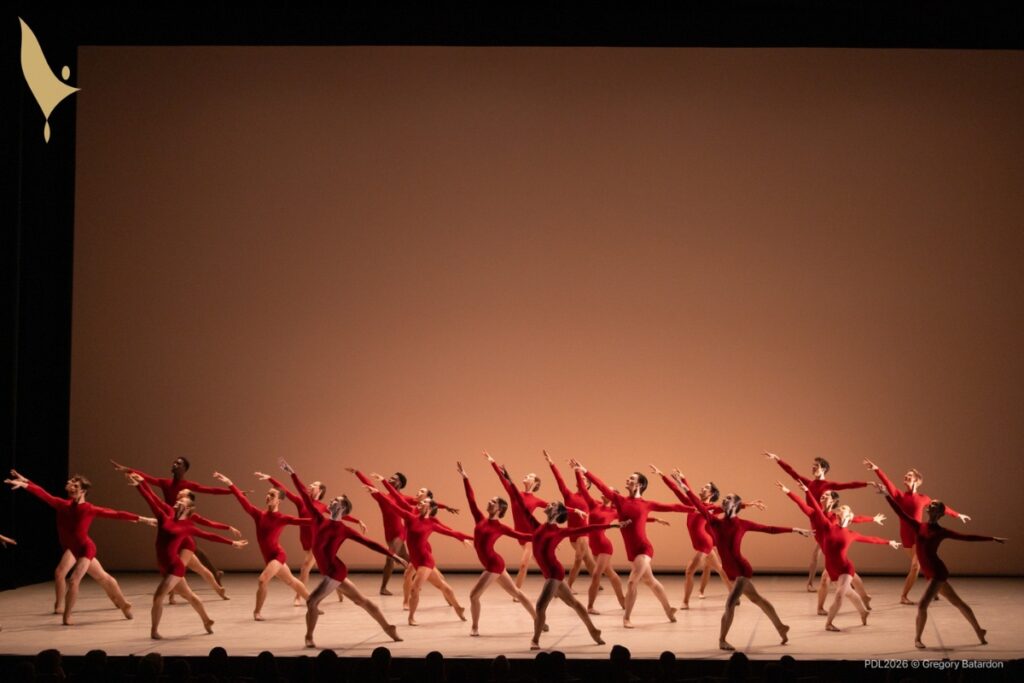 The group of dancers pose in tendu back with their left legs extended and right legs bent. Their arms are outstretched, right arm up and left arm side. They are dressed in red long-sleeve unitards against a light orange background.