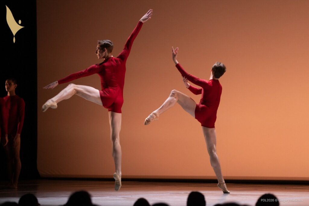 Henry and a male dancer dance in edwaard's 'the gathering. ' henry, left, jumps with his left leg in attitude side with his ride leg extended to the floor. His arms are extended, right arm up and left arm in line with his left leg. The partner is to his right, with his left leg in a matching attitude side and right leg on releve. His arms are bent upwards to the side, mirroring his leg, and he looks upstage away from henry.