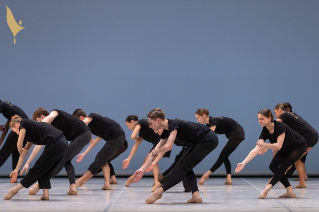 Students dressed in black tights and black t-shirts pose on stage in rehearsal. They bend their right legs with their left legs tendu front croise. Henry is front and centre of the group.