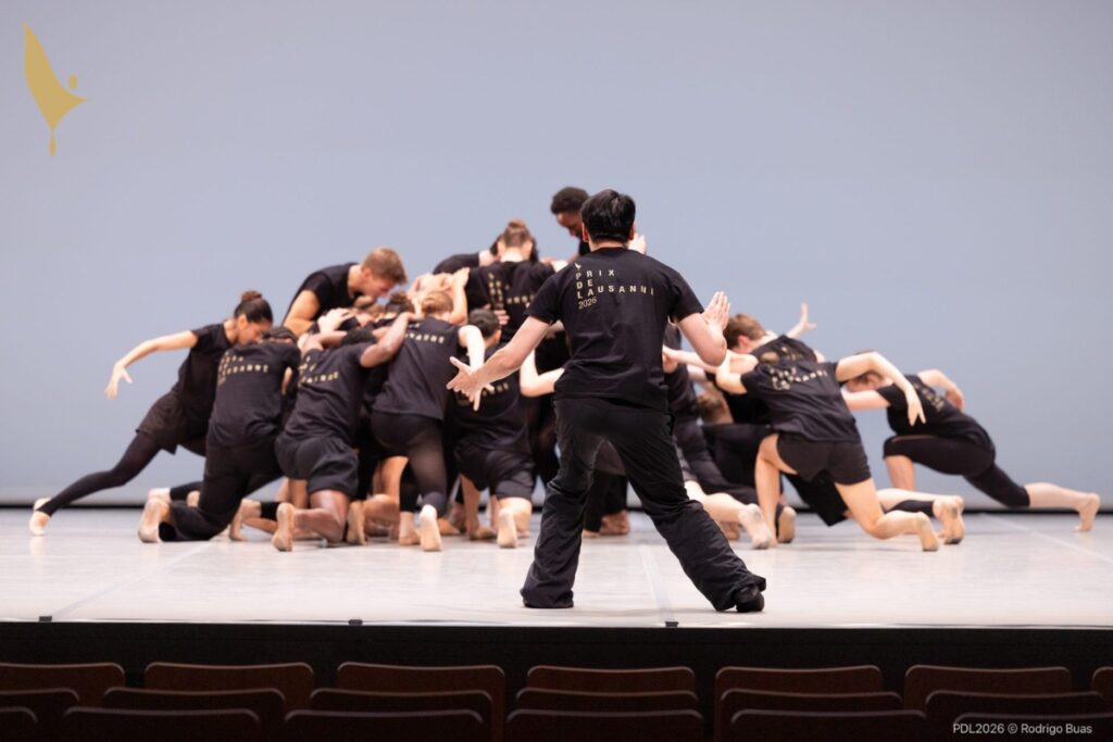 The group of dancers are dressed in black tights and black prix de lausanne t-shirts. Edwaard faces the group up stage, his back to the camera, and squats to mimic the group pose, with all the dancers huddled in a pile.