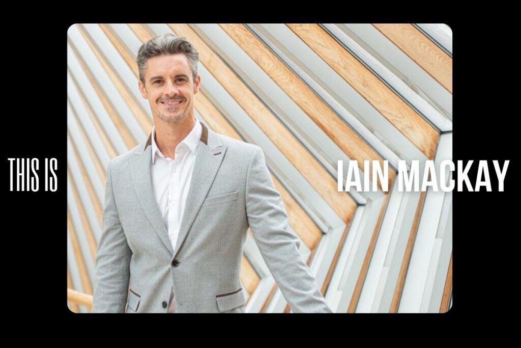 Iain mackay in a white shirt and grey blazer, standing on the bridge of aspiration smiling.