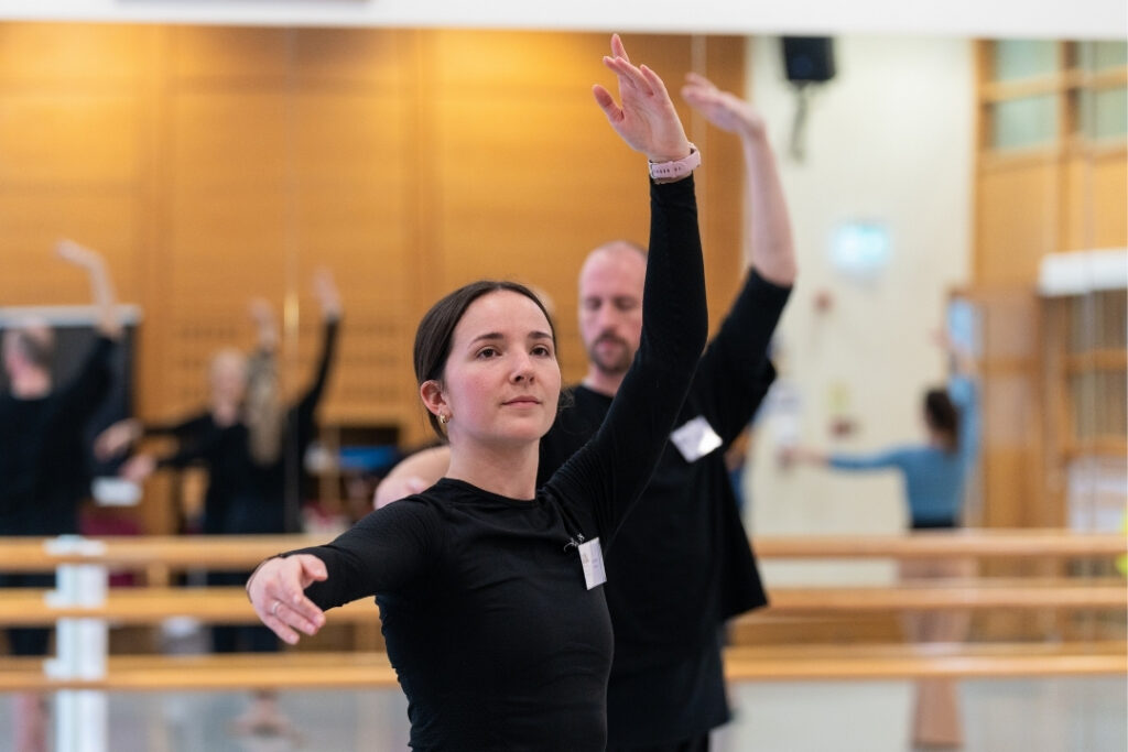Lucy stands with her left arm in fifth and right arm to the side in second in the middle of a ballet class. She wears a black long-sleeved shirt with her hair in a low bun.