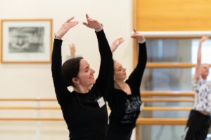 Lucy pictured in a black long-sleeve shirt with her arms up in fifth position. She smiles with her eyes closed. A teacher in the background in a black shirt mirrors her pose.
