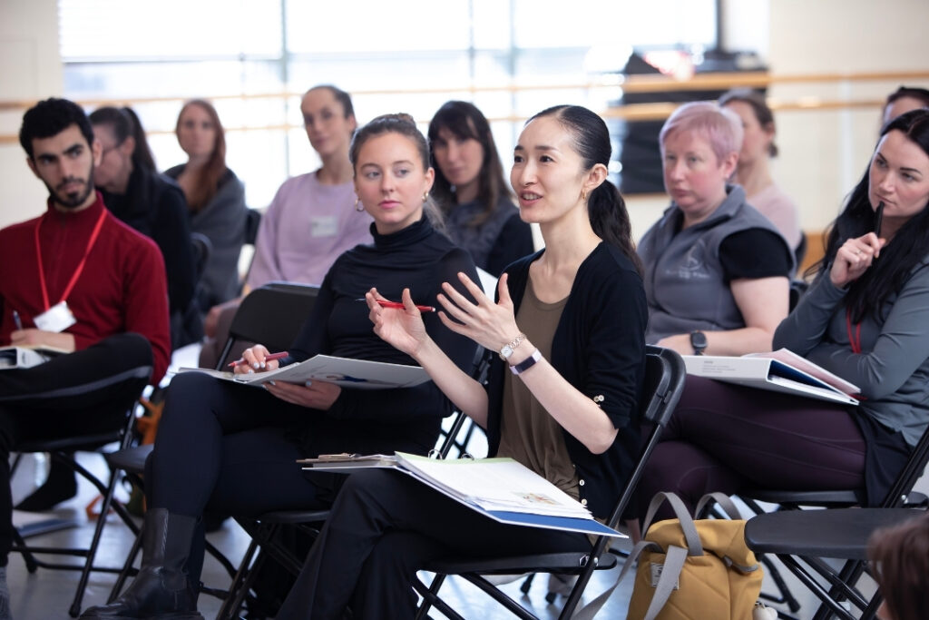 A group of teachers sit in chairs during an inspire seminar. One teacher in the foreground is talking. Her hair is in a low ponytail, and she wears loose-fitting dark coloured clothing. An open notebook sits in her lap.
