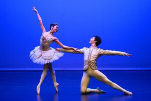 Sophia and julian pose in grand pas classique. Sophia is on pointe in fourth position croise to stage right looking back at julian. Julian kneels next to her with his left leg straight and back. They wear white costumes.