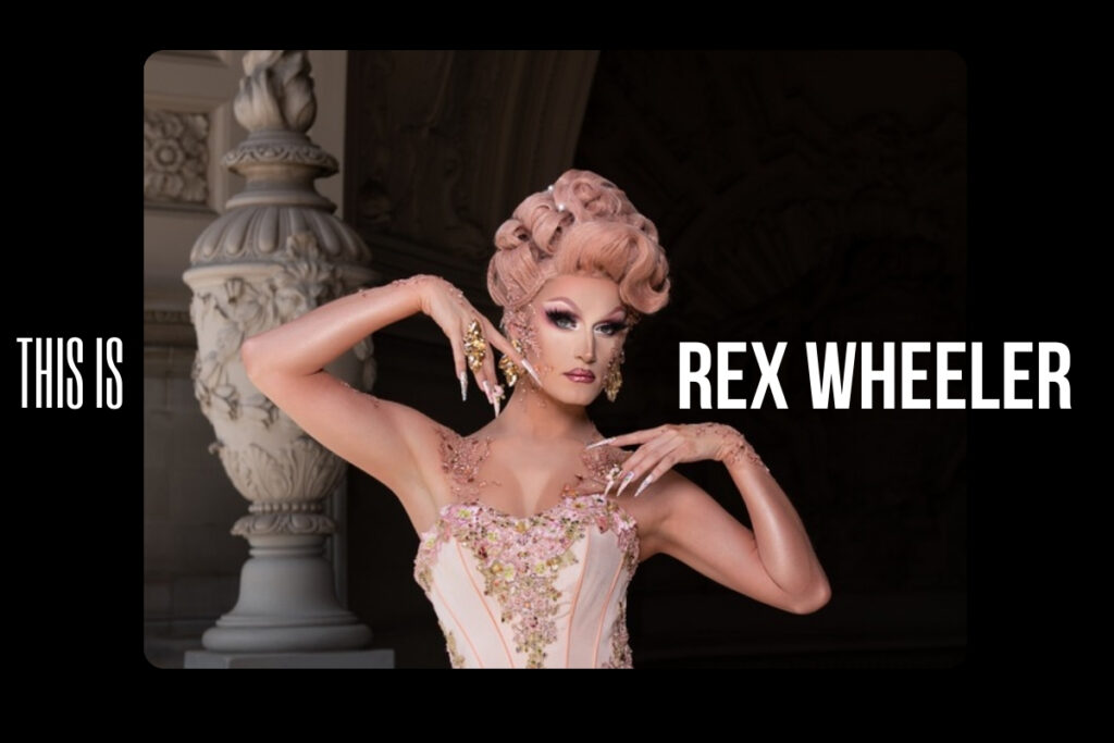 Rex wheeler as lady camden, wearing a light pink tutu and matching pink wig in an elegant updo. She looks into the camera intensely without a smile, her hands framing her face.