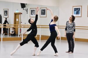 Adesola stands on the right with her head tilted to the side, watching two dancers rehearse. She wear a grey patterned shirt, black trousers and brown ballet shoes. The dancers face each other. The lady on the left plies on her left leg with her right leg tendu back. Her left arm is down to the side, and she looks up to her right arm, raised on a diagonal. The second lady, to her right, tendus her left leg towards her and plies her right leg. She also gazes up towards her right arm, raised in a diagonal. Her left arm is held to the side. They wear black trousers. The first lady wears a long-sleeved black top. The second wears a navy cap sleeve leotard.