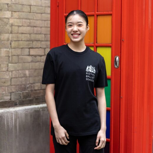 Product image of 'This is 100 years t-shirt' in black. Worn by a Royal Ballet School student in Upper School, Covent Garden. A collection of merchandise in celebration of 100 years of The Royal Ballet School.