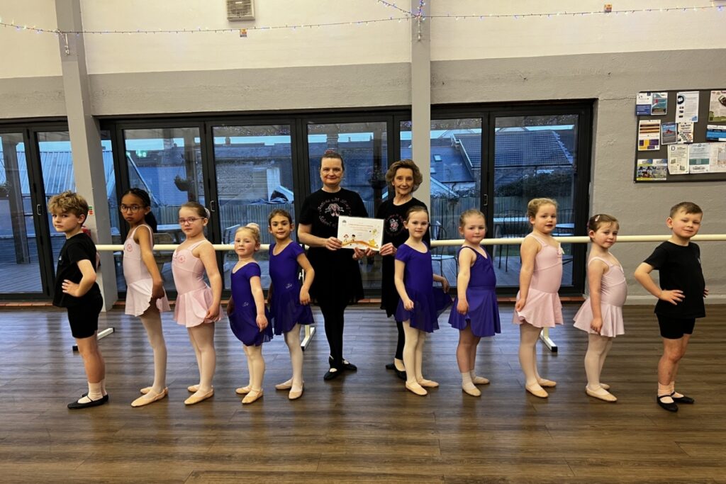 Anthea and alicia king stand with their dance students, who are dressed in cobalt blue and light pink leotards and skirts with pink tights and ballet shoes.