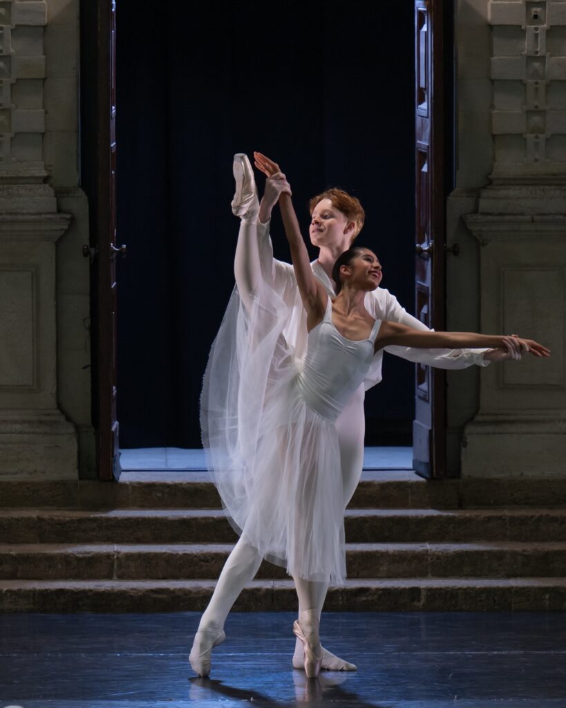 Harry and maria are dressed in white costumes against a dark background at opera holland park. Harry is in a loose fitting long-sleeved white shirt and white tights. Maria is in a white leotard with tank sleeves and a long white tulle skirt. Her right leg is in a developpe with her right arm parallel to it, being held by harry. She is standing on pointe on her left leg. Harry stands behind her holding her arms with his right leg pointed in tendu to the side.