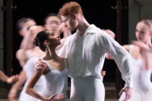Harry and maria hold each other with one hand and gaze into each other's eyes in front of a long-exposure background of dancers moving behind them.