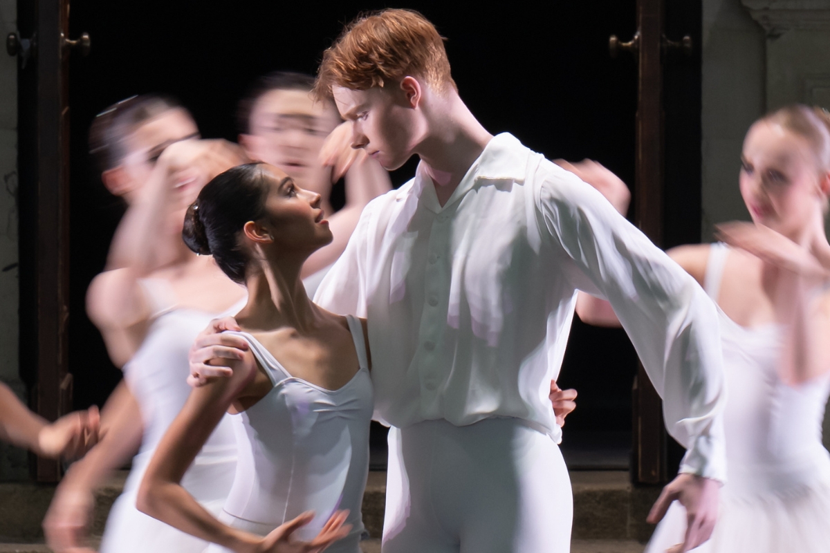 Harry and maria hold each other with one hand and gaze into each other's eyes in front of a long-exposure background of dancers moving behind them.