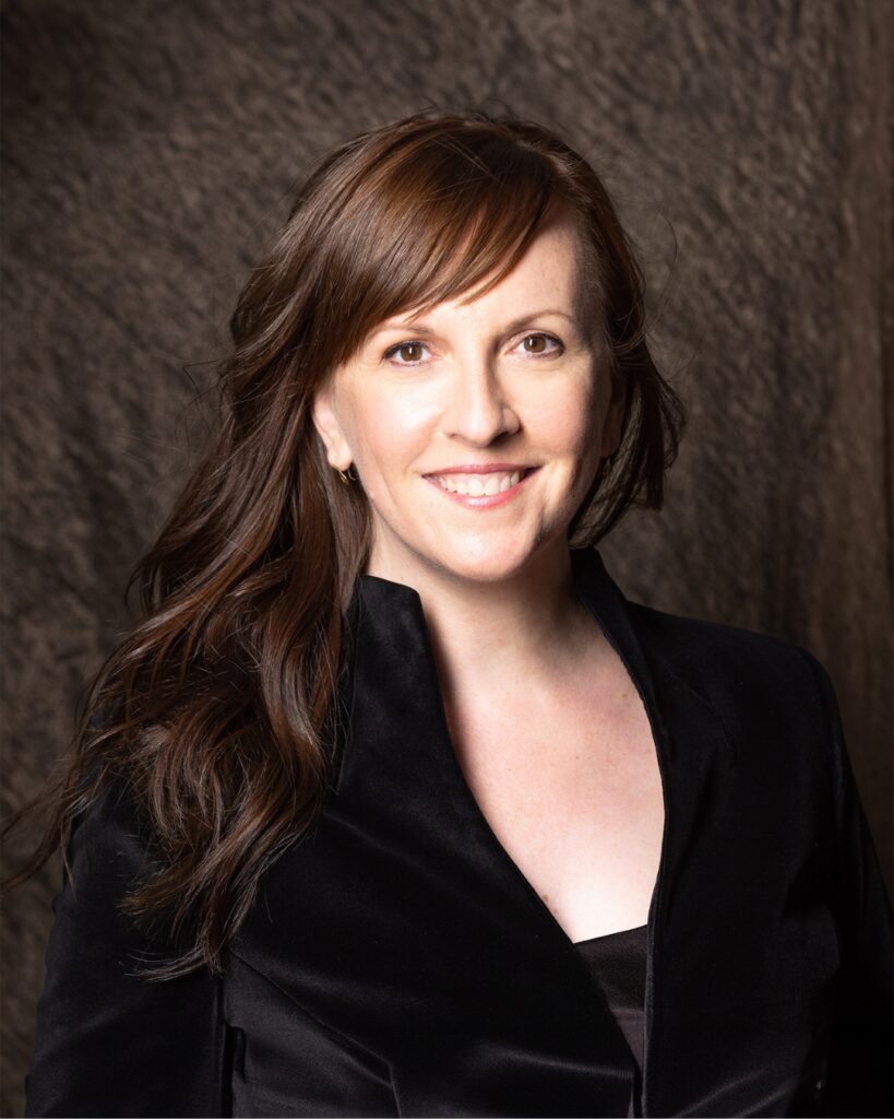 Jessica wears a black v-neck shirt and smiles in front of a textured brown background. Her brown hair is lightly waved and she has bangs swept to the side.