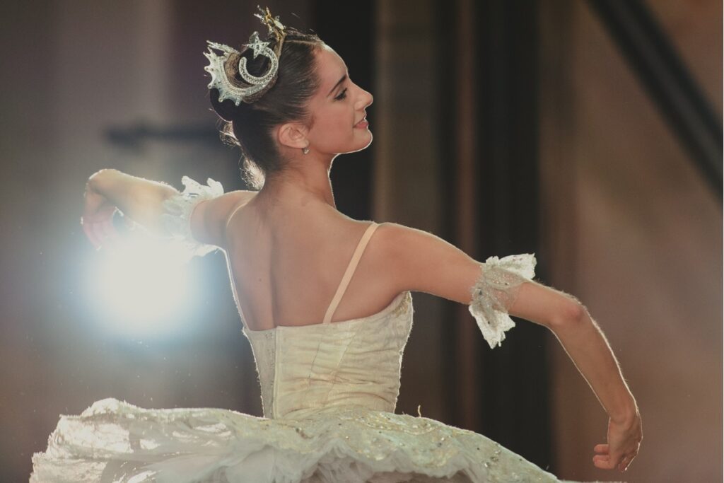 A dancer is on stage in an aurora costume: a white tutu with arm ruffles and a swirling white tiara. She smiles at the audience. Her right arm is bent in front of her with her left arm to the side. The camera is slightly behind her, facing her right side.