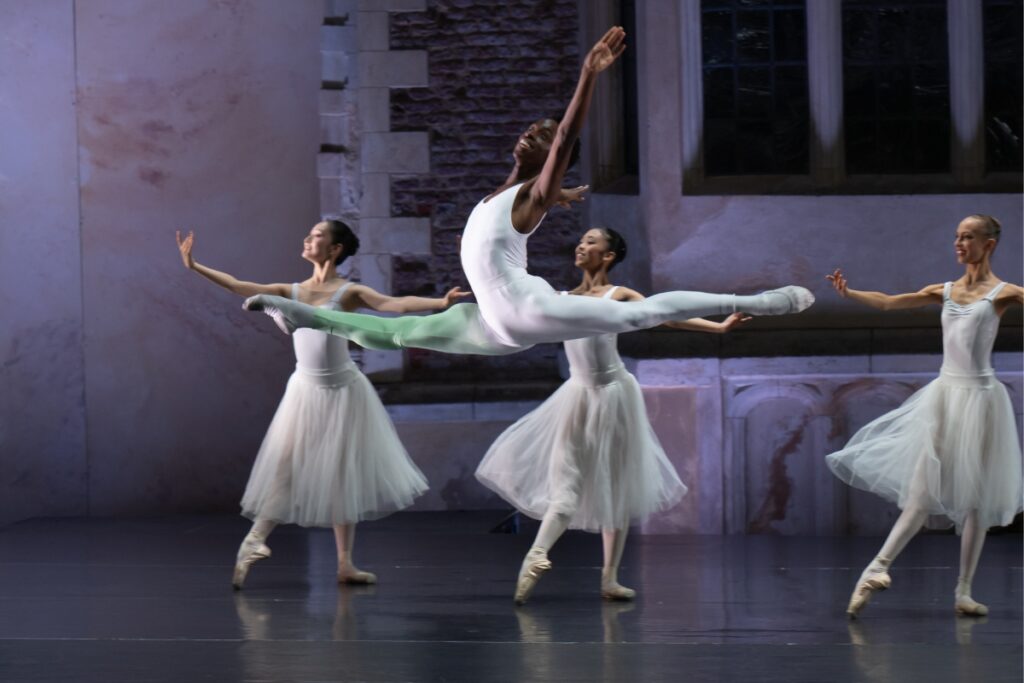 A male dancer jumps mid grand jete with his left arm raised. His right leg is extended front and has a pastel green wash of colour from his ankle to his thigh. Three female dancers pose behind him in a tendu front with their right leg and their right arm extended front with a gentle curve and palm up. Their left arms are held to the side, also palm up. All dancers brightly smile.