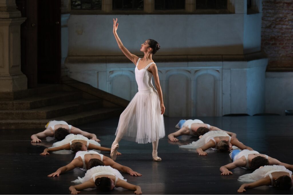 A female dancer poses with her right arm raised and gently curved in front of her face. She stands between two rows of dancers lying on the floor with their palms flat next to their heads.