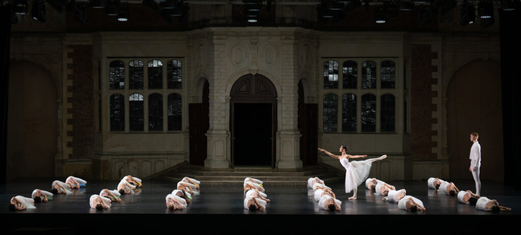 Dancers dressed in white lie on the stage at opera holland park in rows. Maria stands between the rows in an arabesque, facing stage right. Harry stands stage left looking at her.