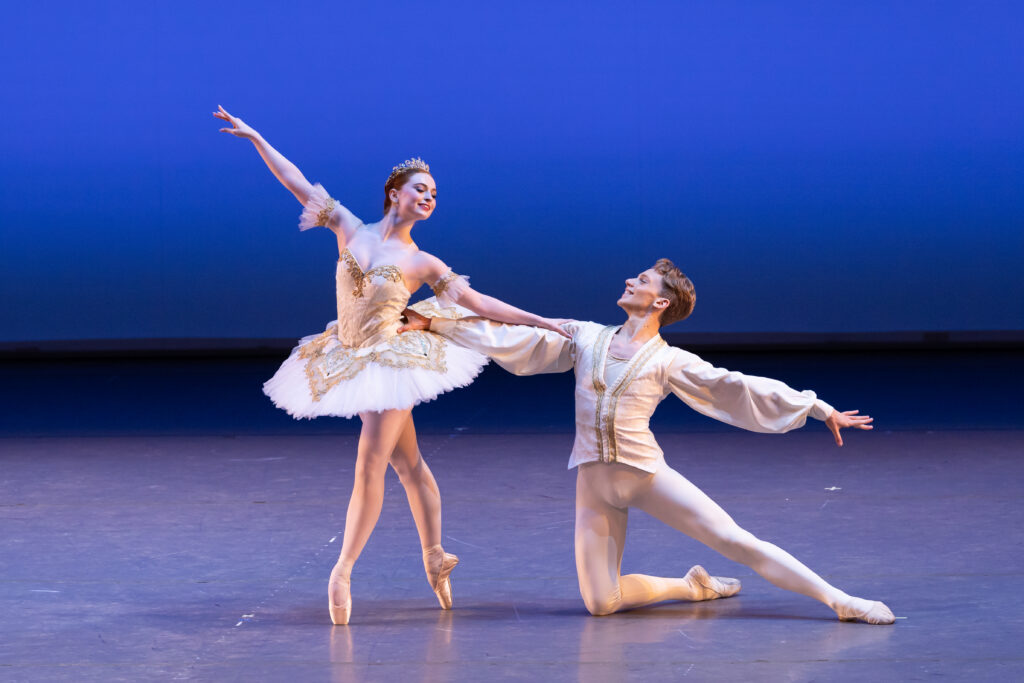 Samantha stands in a fourth position on pointe croise with her left leg front and downstage. Her right arm is raised on a diagonal, and her left is on a downwards diagonal towards ivan. She wears a glittery white tutu and headpiece. Ivan wears a white tunic and tights. He kneels on his right leg with his left leg extended to the side. His right arm holds samantha's waist while his left is also on a downwards diagonal.