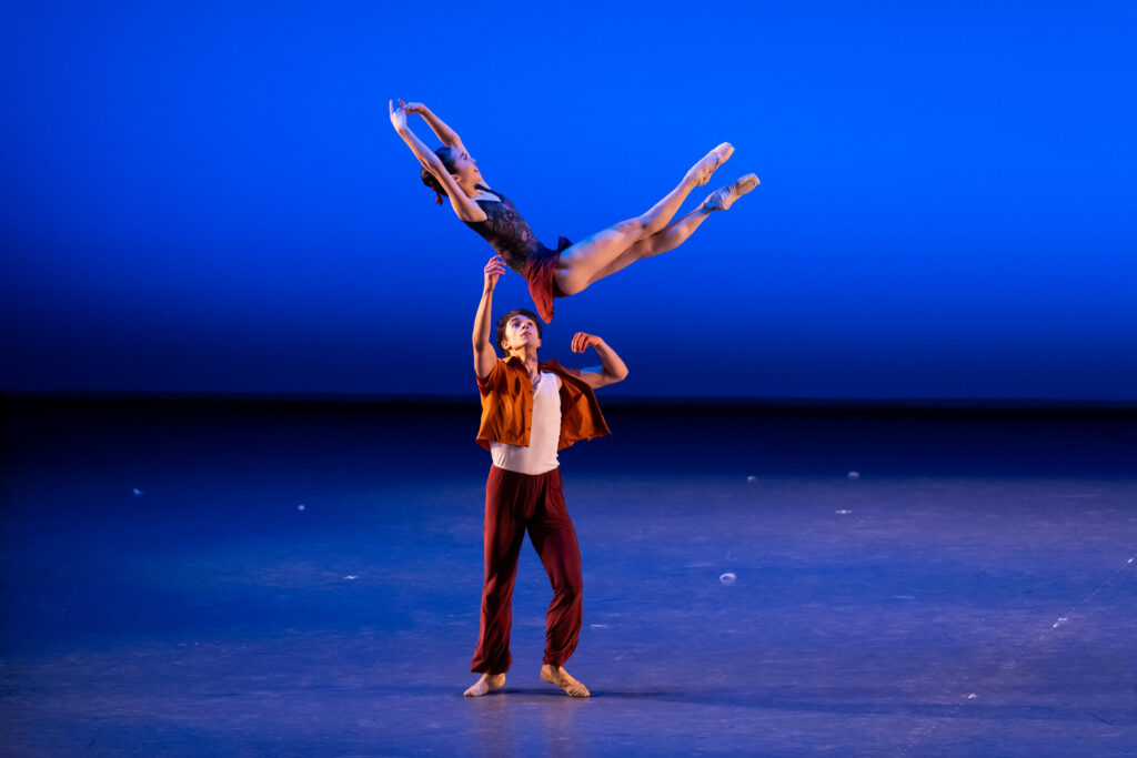 Against a bright blue backdrop, millan tosses nanoha into the air. Nanoha's body is at a 90 degree angle, with her legs crossed in fifth position in front of her on a diagonal to the upper right. Her torso is on a diagonal to the left of the image with her arms in fifth. Millan is below her, arms raised and poised to catch her.