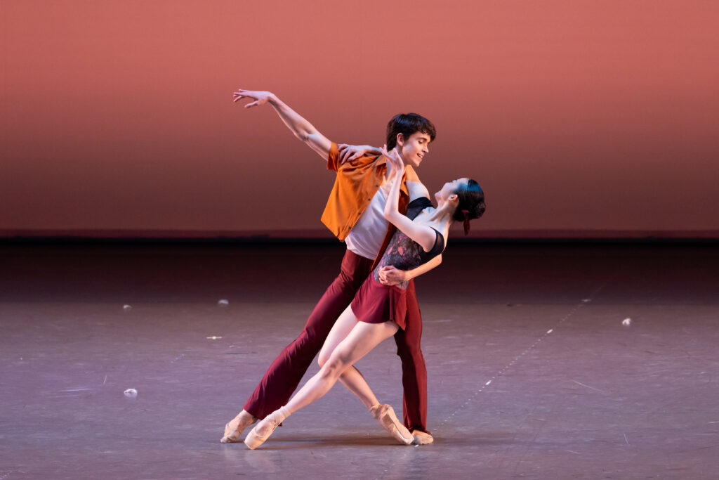 Millan and nanoha pose centre stage against a warm red-orange backdrop. Millan lunges to the left with his left arm wrapped around nanoha's waist and his right arm raised on a high diagonal. Nanoha leans back into his arm and cups his jaw gently with her left hand. Her left leg is extended to the front, parallel to millan's extended right leg. Her right leg is bent, propping her weight on her back foot.