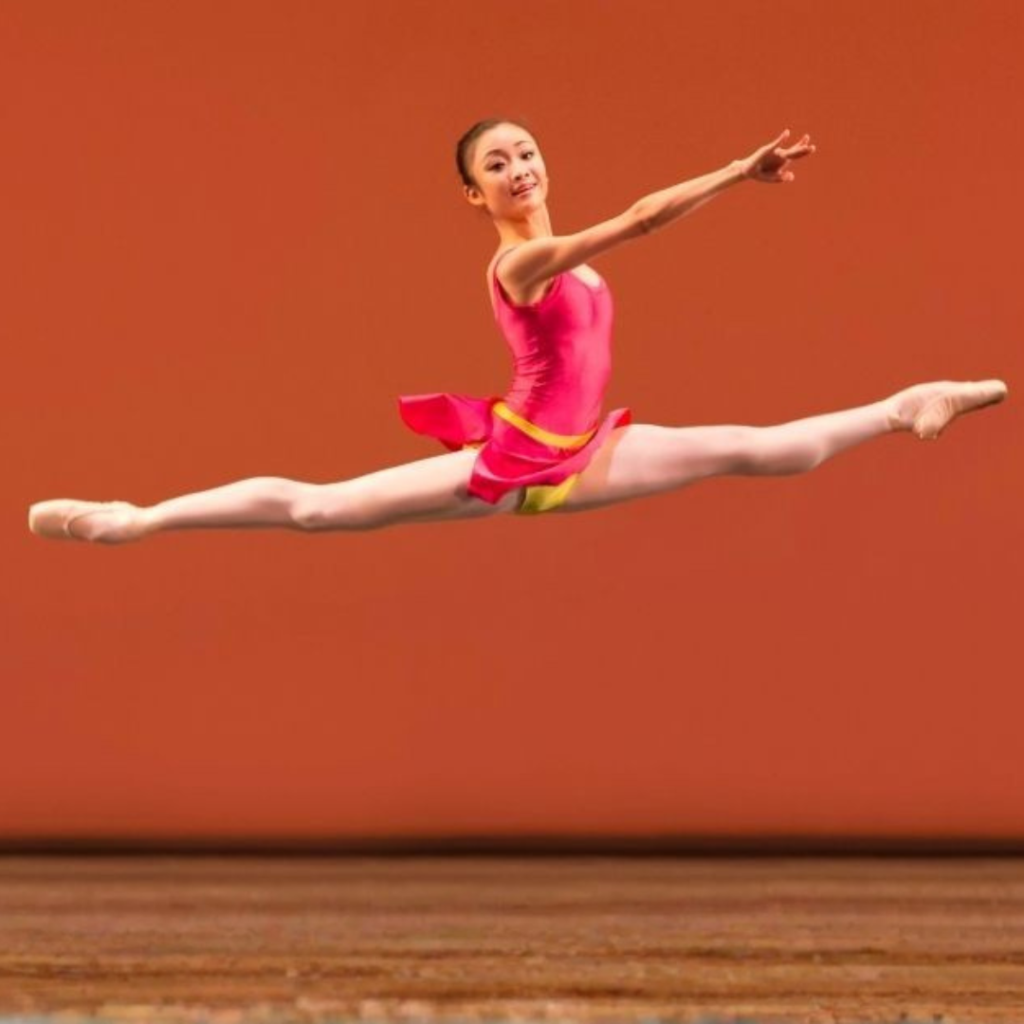 Yaoqian leaping through the air in a pink leotard in front of an orange backdrop.