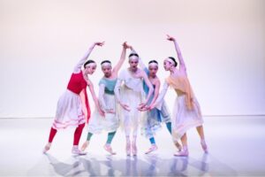 Dancers in red, green, white, blue and yellow tunics with light shimmery headbands pose in a semicircle. Their arms are curved above their heads as they lean towards each other. Their gazes are down towards the centre ground.
