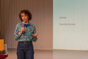 Dr adesola holds the microphone in front of her presentation slides. She wears a blue shirt with a colourful print and jeans.