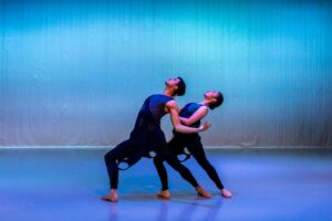 Two dancers lunge with their right legs forward, arching their backs and gazing to the ceiling. The dancers legs overlap, and the dancer in the back holds the arms of the dancer in front, whose arms are bent to the back, palms up.